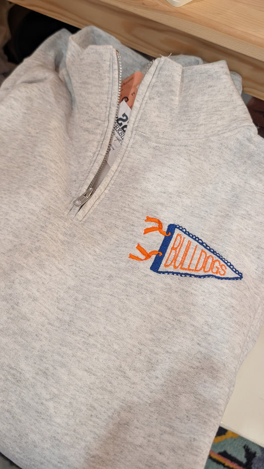 A light heather gray quarter-zip sweatshirt with an embroidered orange and blue 'Allugs' pennant on the left chest, shown folded on a wooden surface.