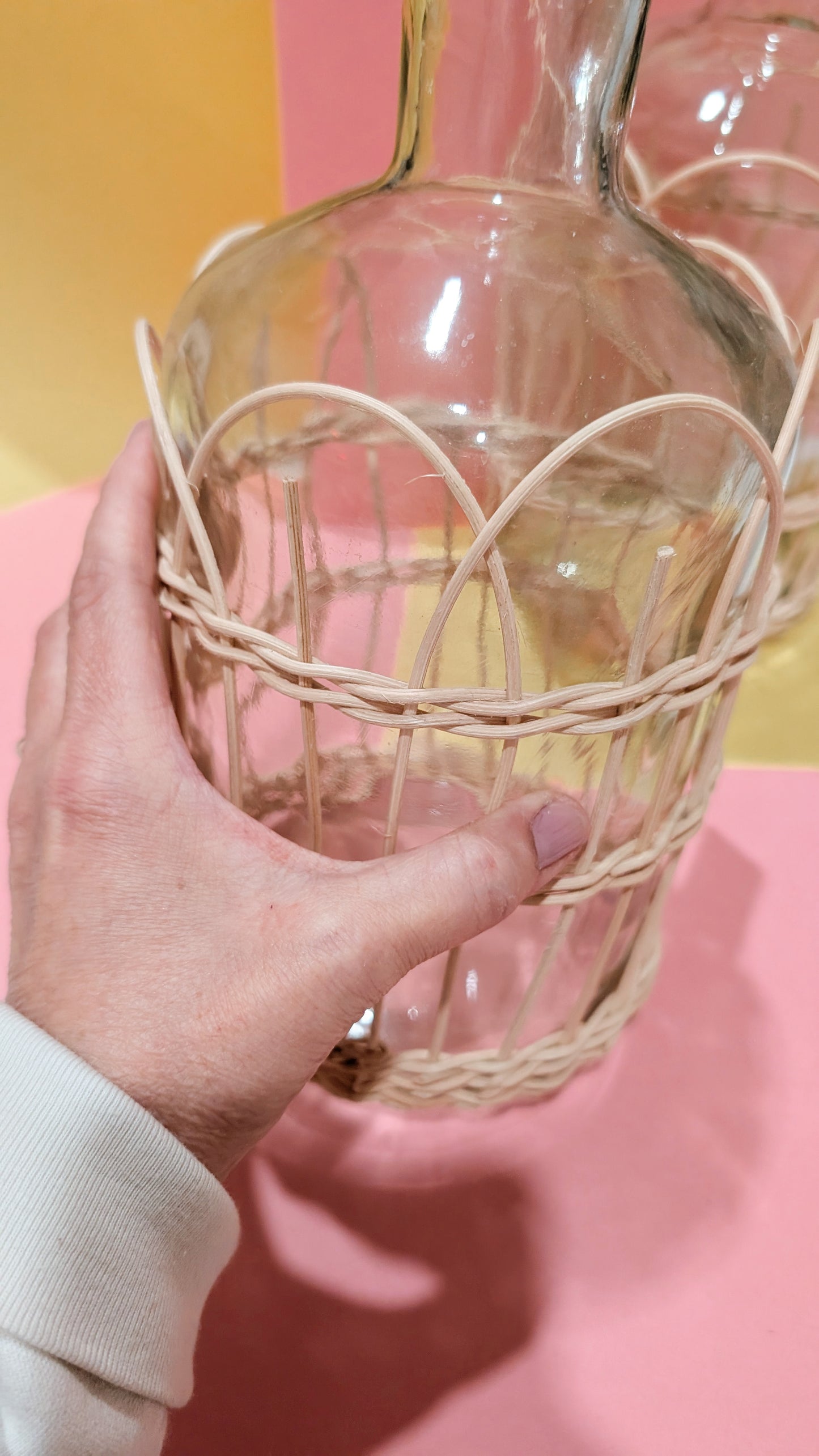 Hand holding a woven basket with a glass bottle inside against a pink background