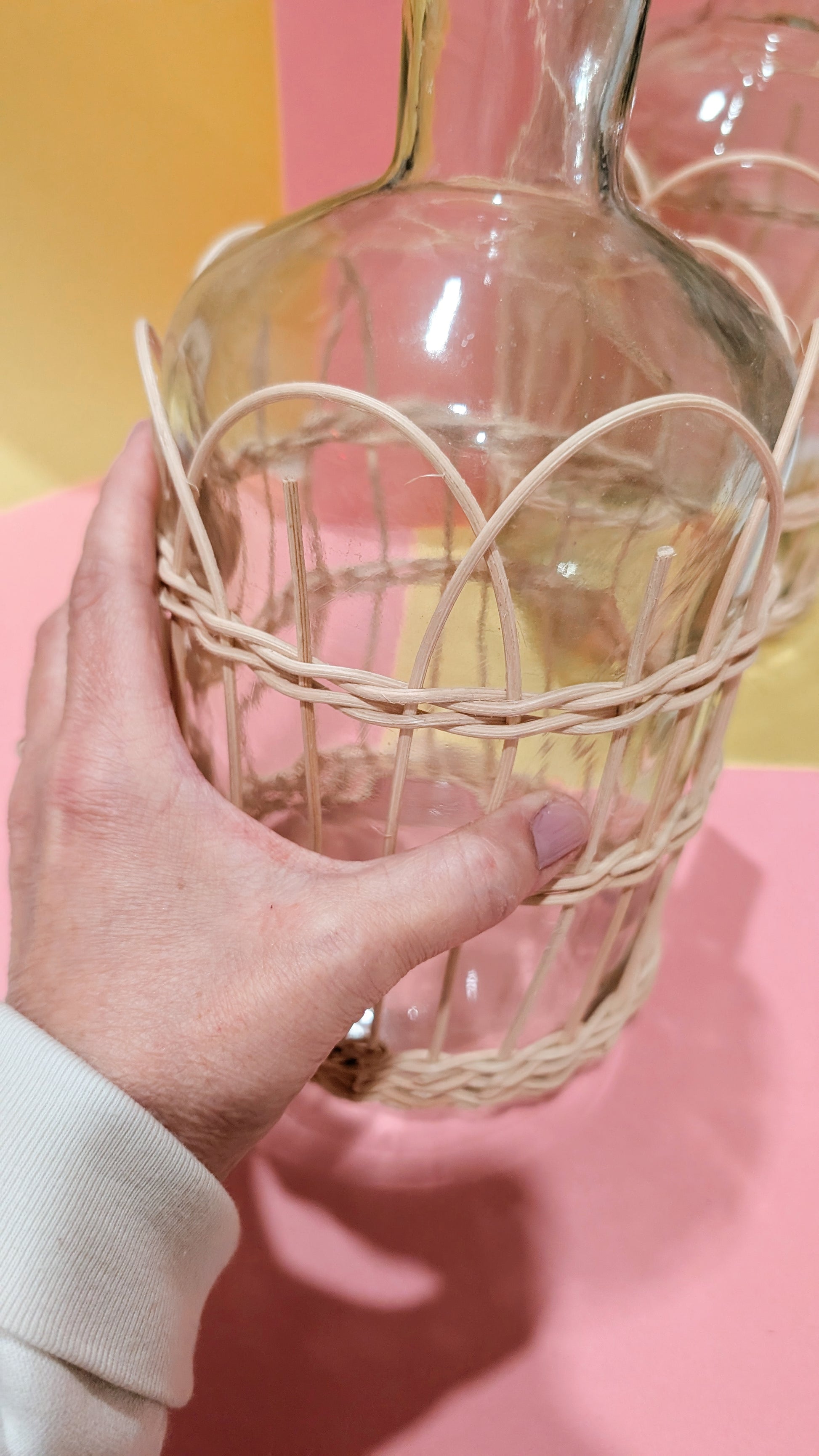 Hand holding a woven basket with a glass bottle inside against a pink background