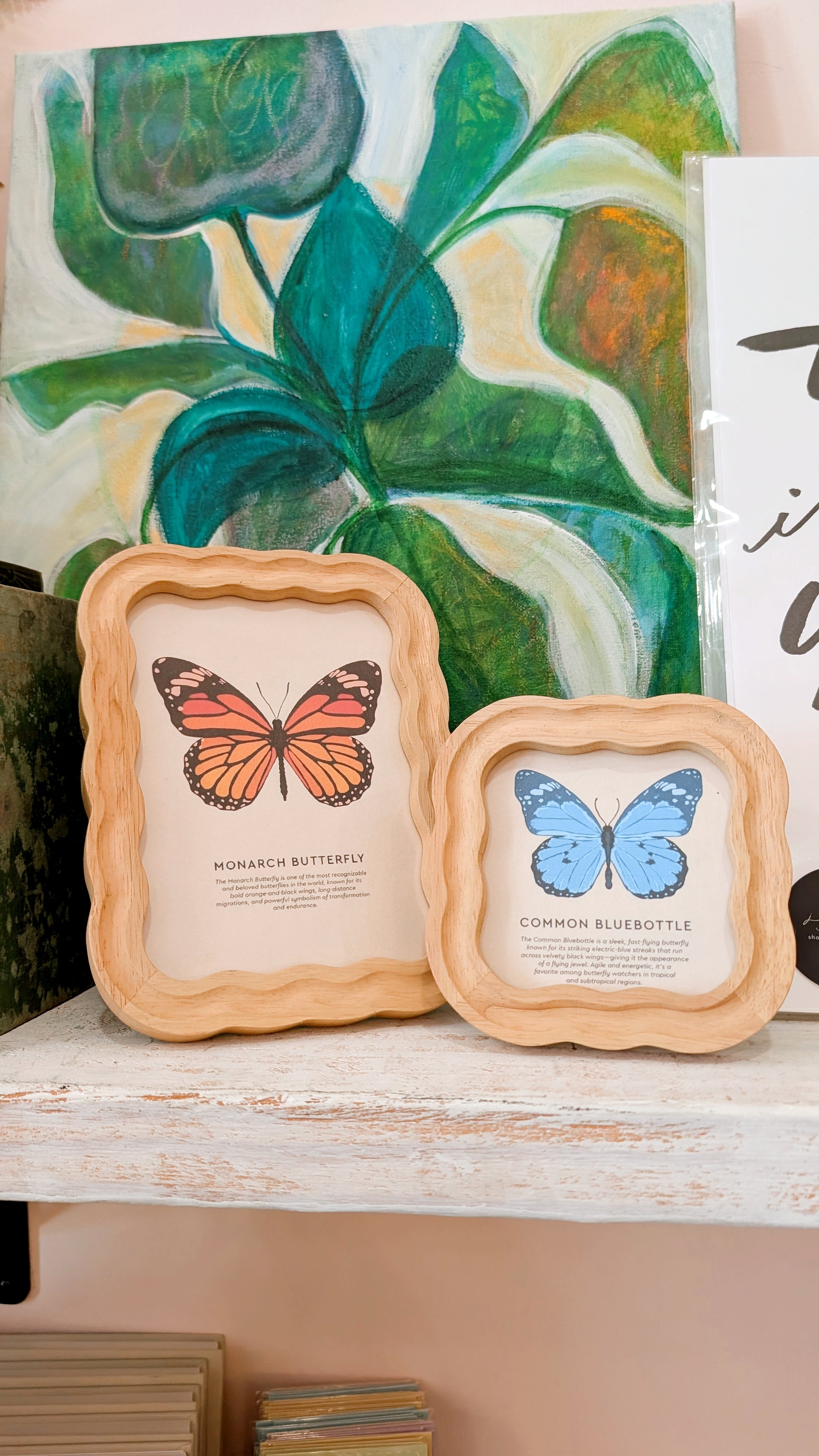 Two framed butterfly illustrations on a shelf with a painting of leaves in the background.