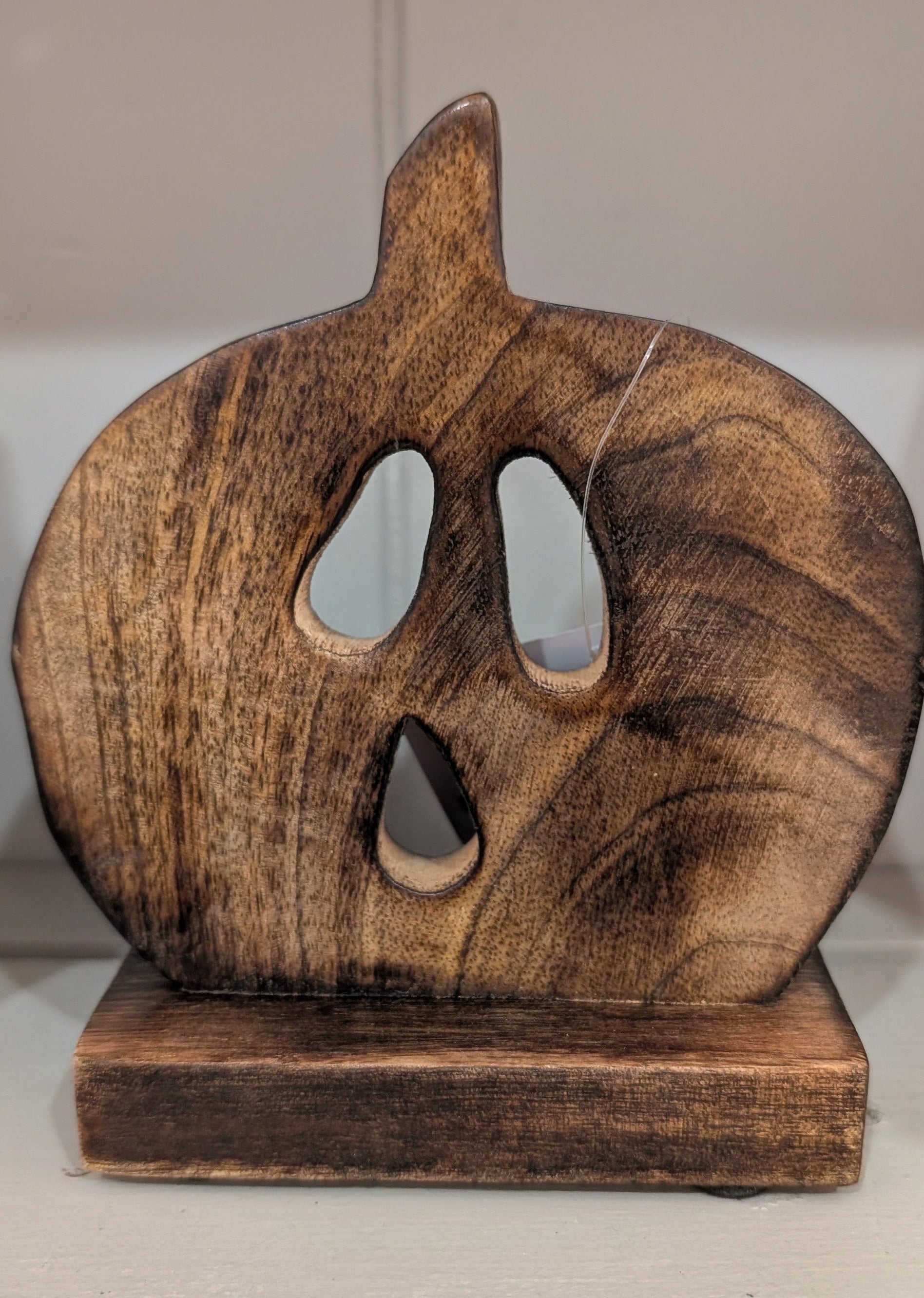 A rustic, handcrafted wooden jack-o'-lantern decoration with carved eyes and mouth, resting on a matching wooden base against a plain indoor background.