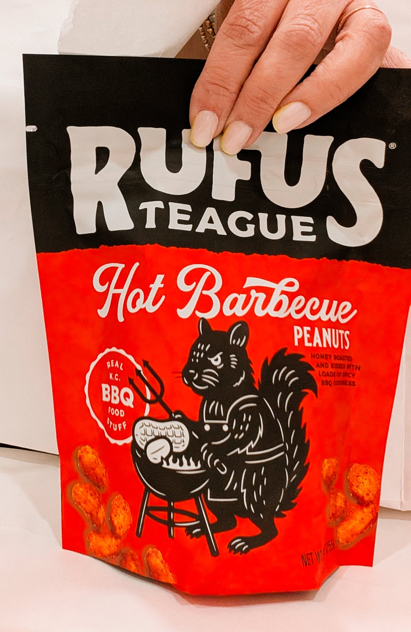 Hand holding a Rufus Teague Hot Barbecue Peanuts package with a black and white illustration of a squirrel on a grill.