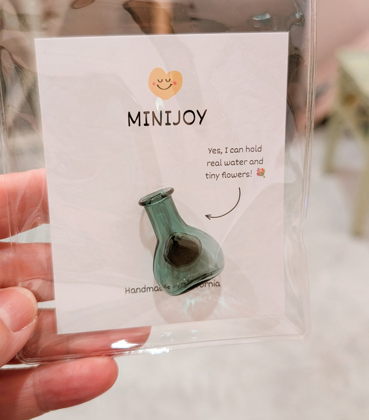 A hand holding a clear plastic bag containing a MiniJoy brand card and a small green glass bottle-shaped charm. The white card features the brand name 'MINIJOY' with a smiley face logo and humorous text.