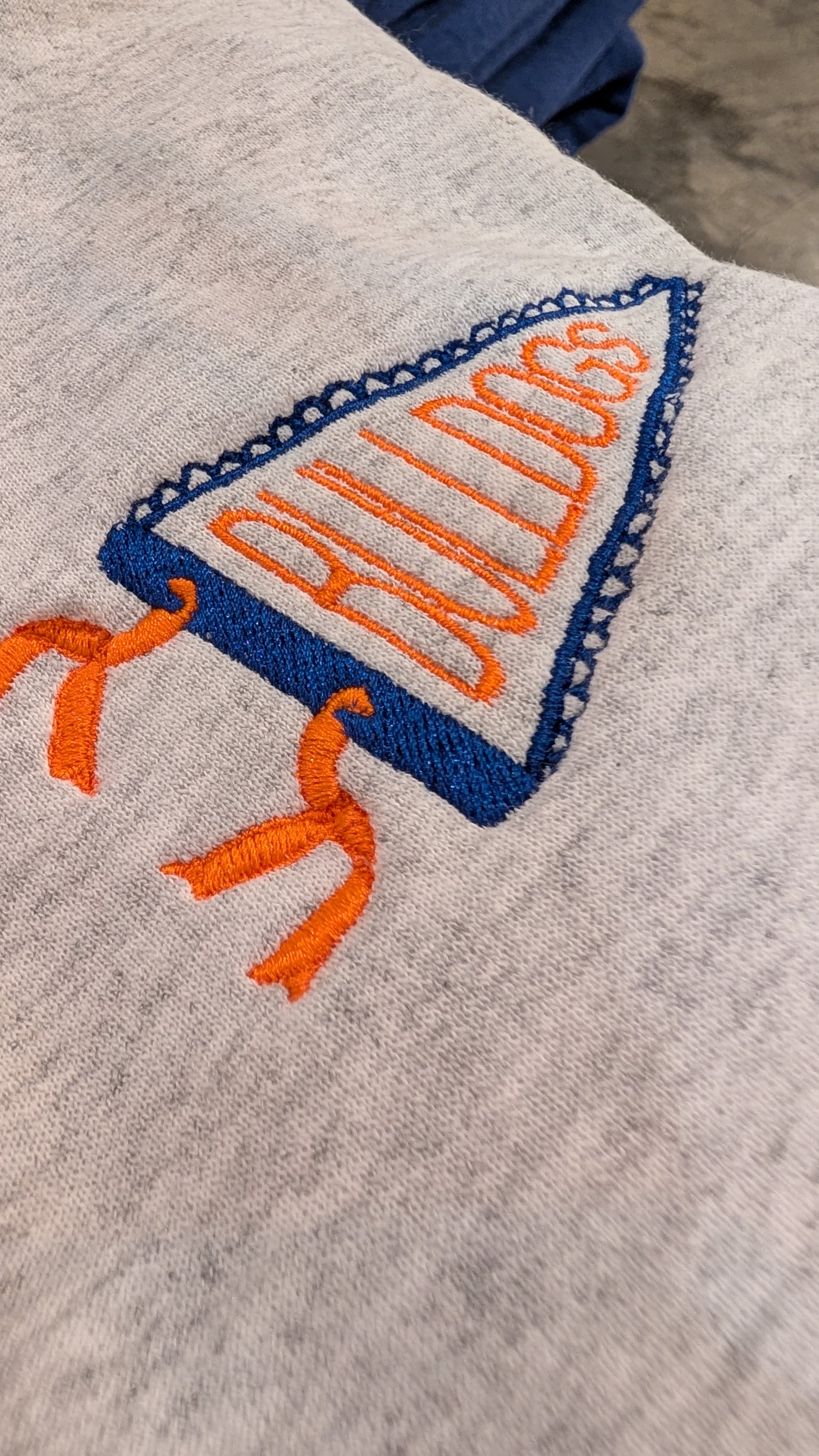 Close-up of a light heather gray fabric item featuring an embroidered blue and orange triangular 'BULLDOGS' design with tassels.