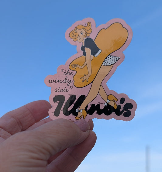 Hand holding a sticker of a woman in a dress with 'Illinois' text against a blue sky.