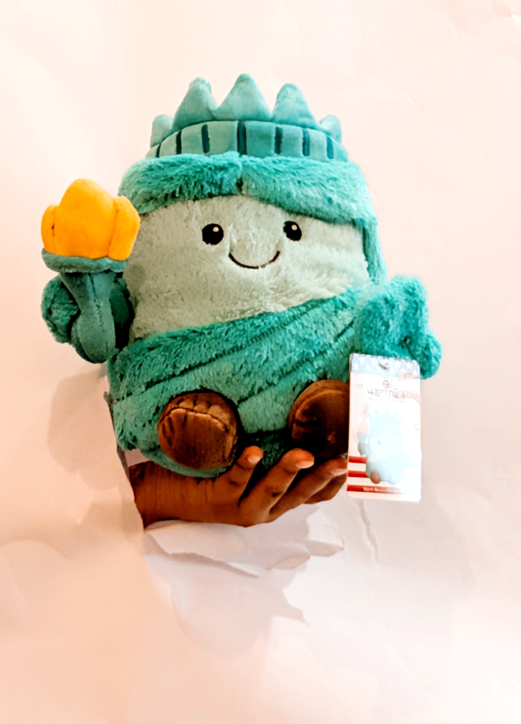 Plush toy resembling the Statue of Liberty held by a hand on a light pink background