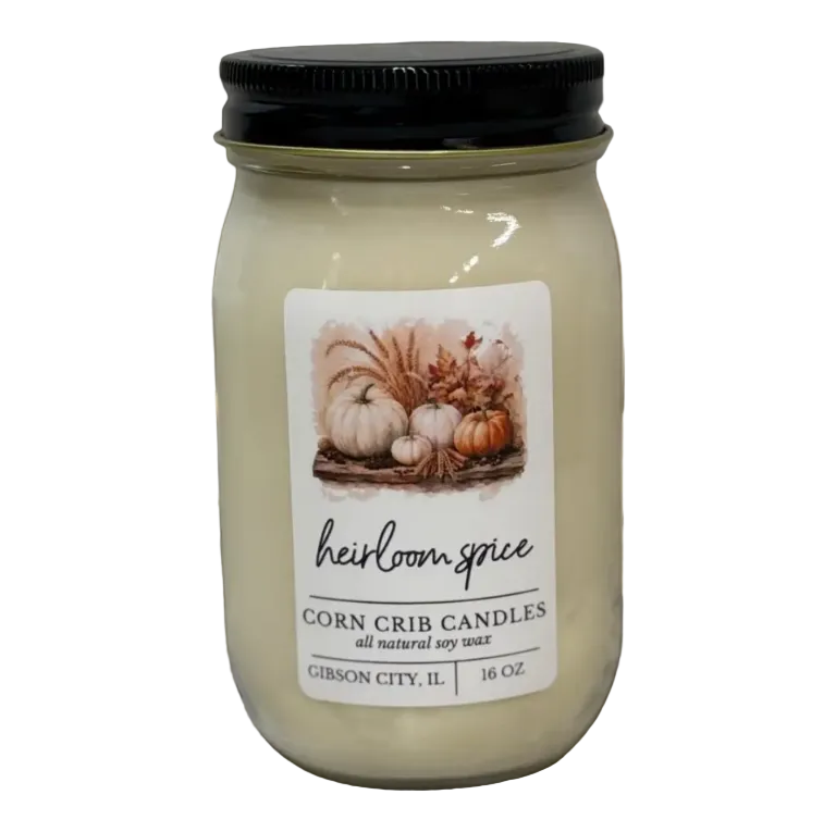 A 16 oz jar candle in a clear glass mason jar with a black lid, featuring a white label with a fall harvest illustration and the brand name 'heirloom spice'. The jar is placed on a rustic wooden table with decorative wheat stalks in the background.