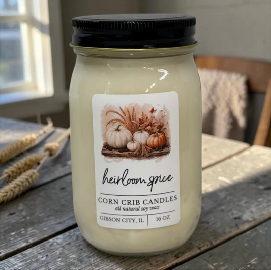 A 16 oz jar candle in a clear glass mason jar with a black lid, featuring a white label with a fall harvest illustration and the brand name 'heirloom spice'. The jar is placed on a rustic wooden table with decorative wheat stalks in the background.