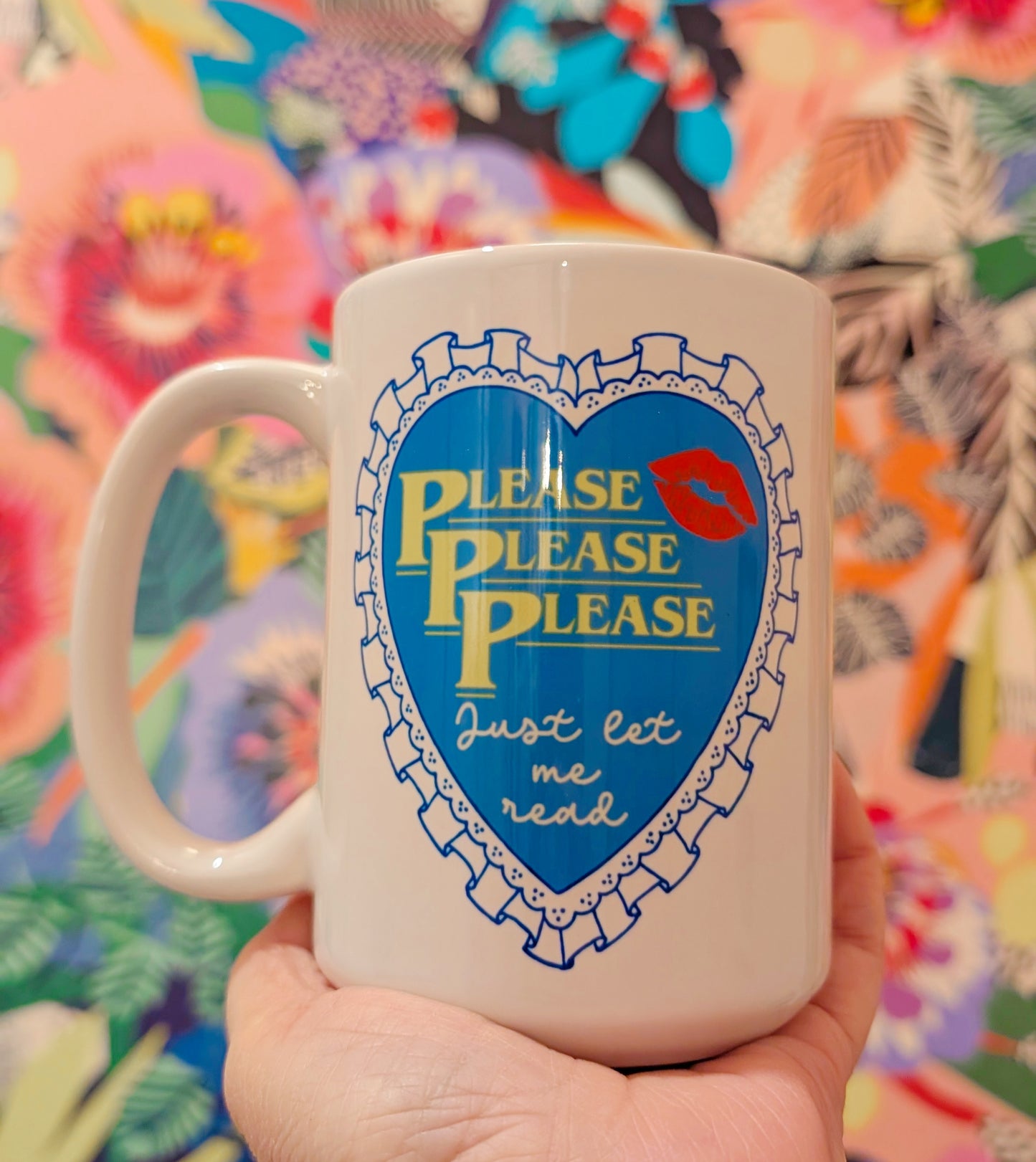 White mug with blue heart design and text held in front of a colorful floral background