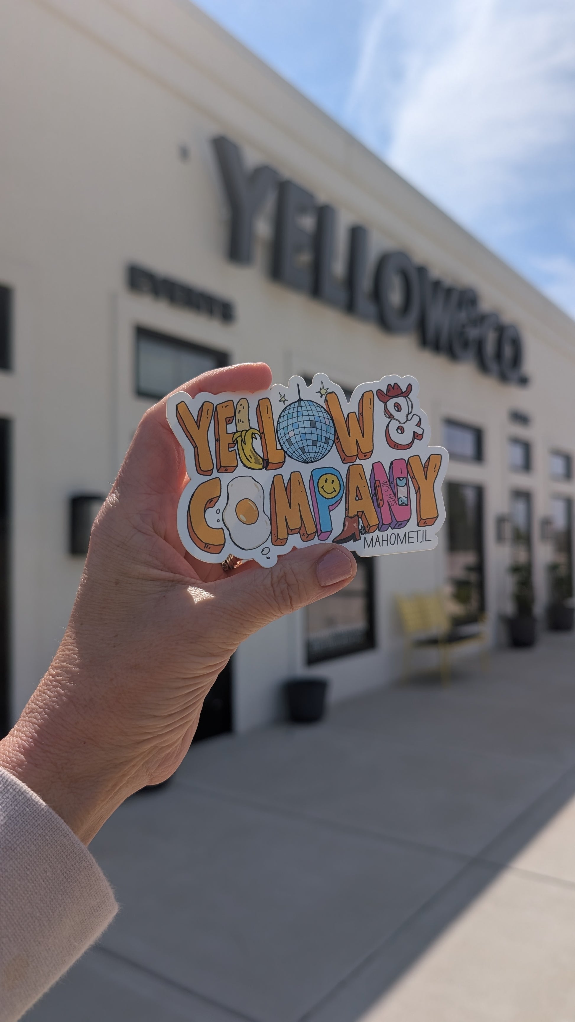 Hand holding a colorful 'Yellow & Company' sticker in front of a building with the same name.