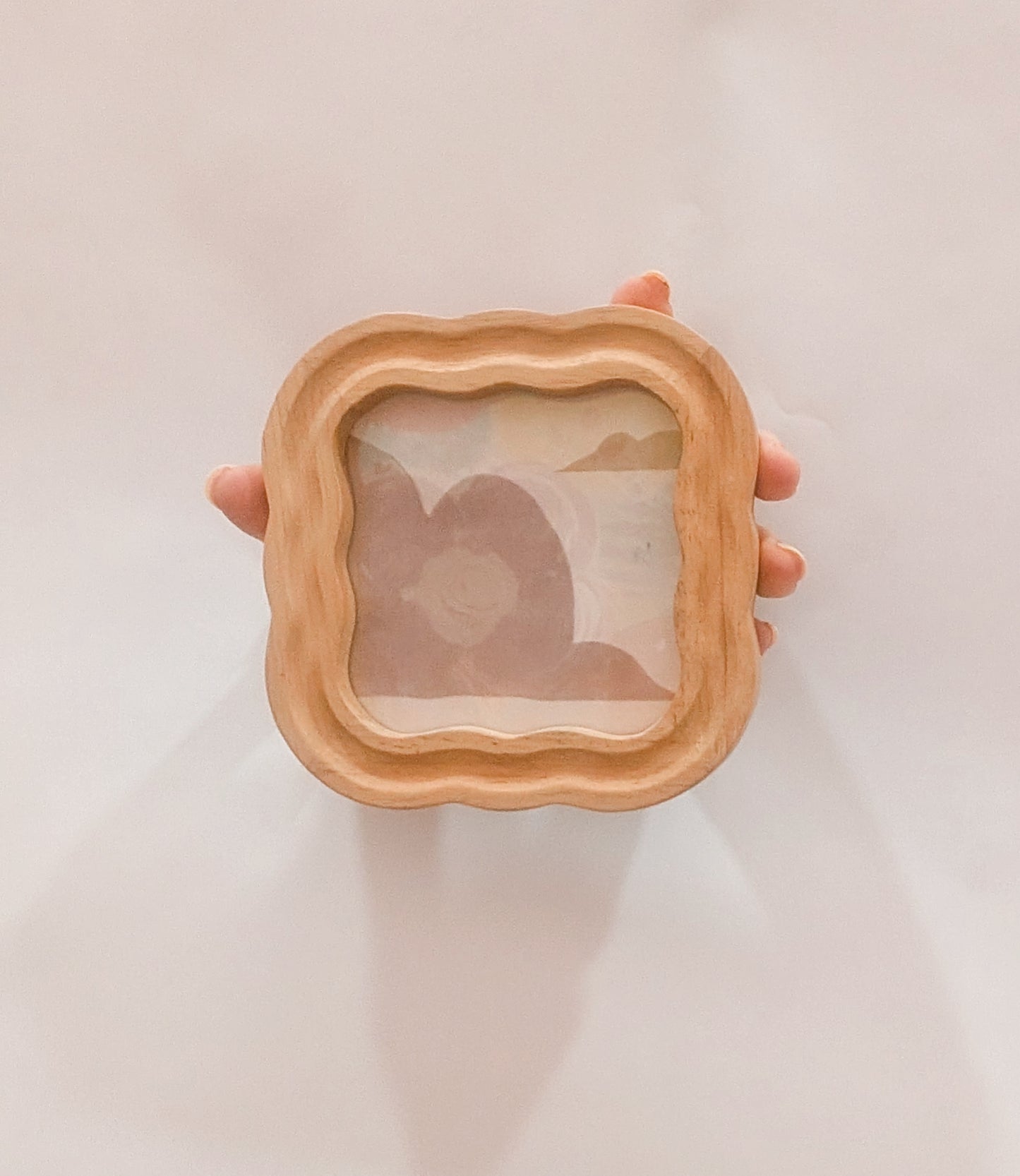 Hand holding a square wooden frame with a scenic view inside on a light background