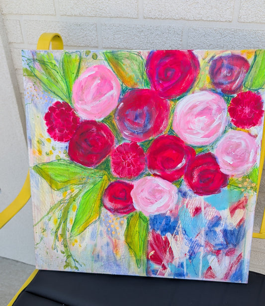 A square canvas painting of a vibrant floral bouquet with pink, red, and white roses and green leaves, displayed on a yellow chair against a light gray brick wall.