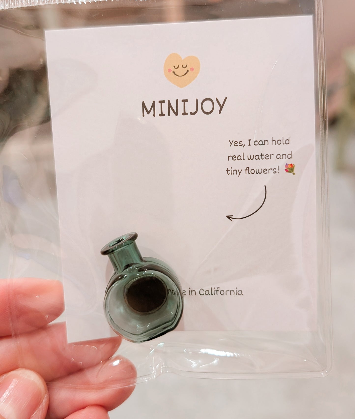 A hand holding a clear plastic bag containing a tiny green glass bottle-shaped accessory attached to a white card printed with the brand name MINIJOY and a small humorous caption.