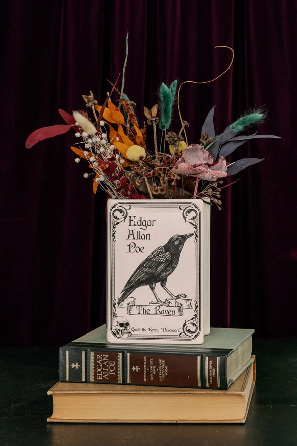 A decorative book-shaped vase featuring Edgar Allan Poe's 'The Raven' cover art, filled with dried flowers and placed on top of two vintage books against a dark burgundy curtain backdrop.