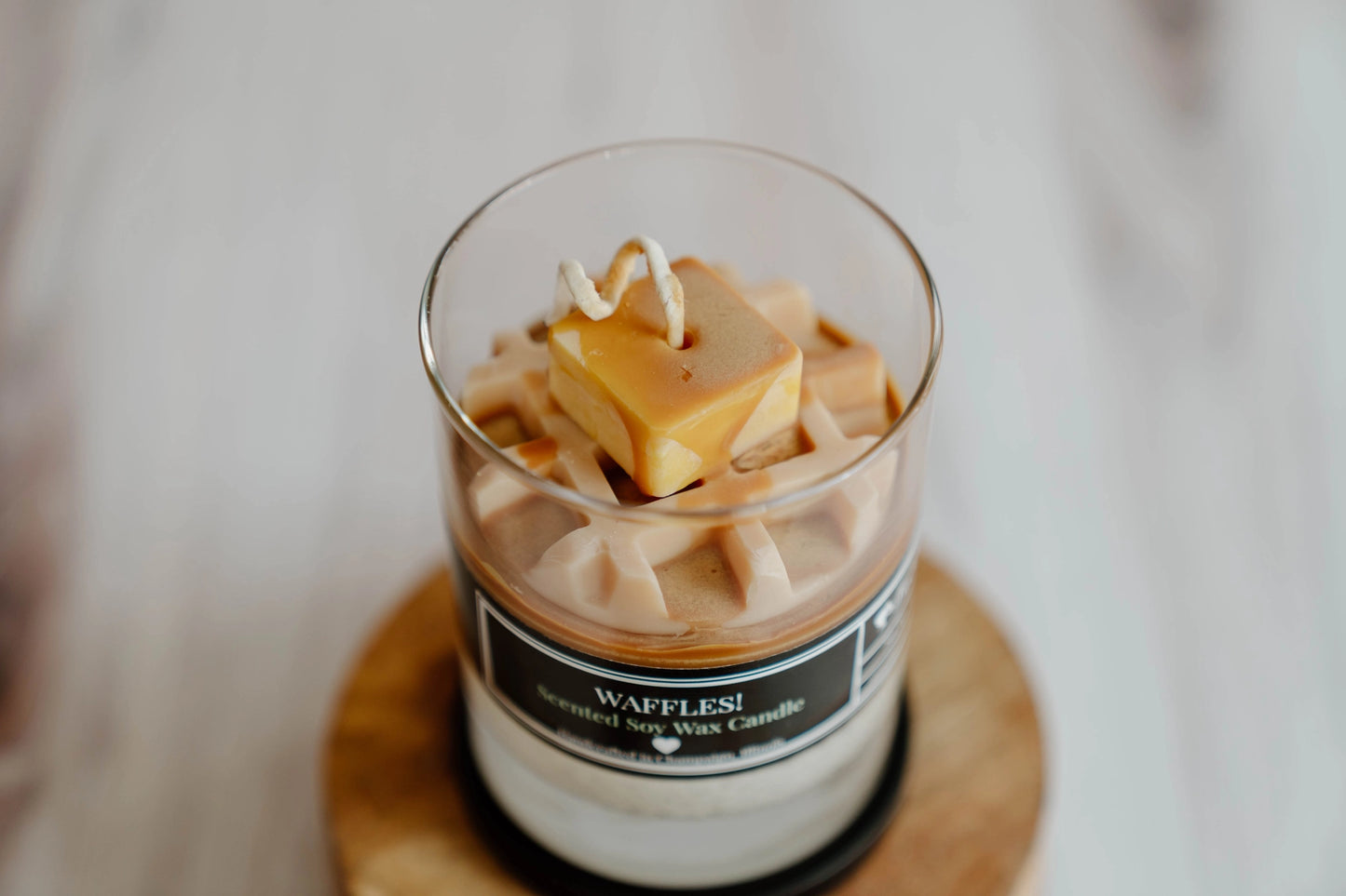 Candle in a glass jar labeled 'Waffles!' with a waffle on top, on a blurred background.